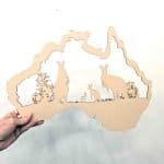 A hand holds a cutout shaped like the map of Australia, made from light brown MDF. Inside the outline are silhouettes of three kangaroos of varying sizes and small bushes, creating a scene that represents Australian wildlife. The background is plain white, highlighting the detailed cutout design.