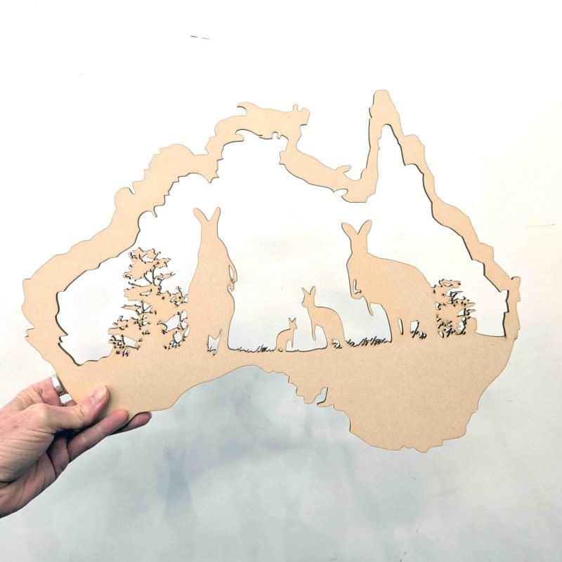 A hand holds a cutout shaped like the map of Australia, made from light brown MDF. Inside the outline are silhouettes of three kangaroos of varying sizes and small bushes, creating a scene that represents Australian wildlife. The background is plain white, highlighting the detailed cutout design.