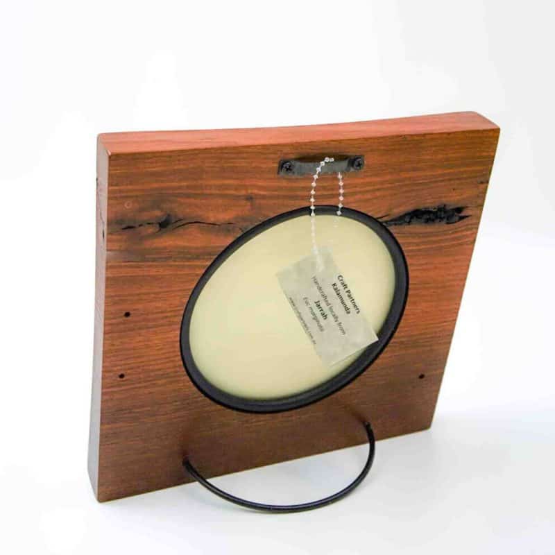 The image shows the back of a handcrafted wooden photograph frame made from Jarrah hardwood. The frame has a rich reddish-brown finish with visible natural grain patterns and dark knots. A black oval photo insert is centered on the back, bordered by a slim frame. A small metal chain with a tag is attached to a hanging bracket near the top, and a curved black metal stand supports the frame from below, allowing it to stand upright. The craftsmanship and texture of the Jarrah wood are clearly visible against a plain white background.