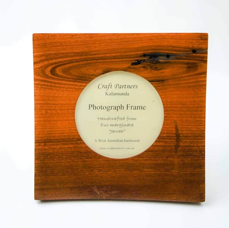 A handcrafted wooden photograph frame made from rich reddish-brown Jarrah hardwood. The square frame features a circular opening in the center displaying a printed label that reads: “Craft Partners Kalamunda – Photograph Frame – Handcrafted from Euc marginata ‘Jarrah’ – A West Australian hardwood.” The wood grain is beautifully visible, with natural variations and darker streaks that highlight the organic texture and craftsmanship of the piece.