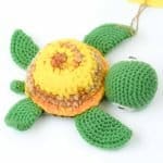 Baby Turtle by Gala Crochet (1) Hand‑crocheted turtle toy made from soft yarn, featuring a round shell in shades of yellow, orange, and light brown, with green crocheted head, flippers, and tail. The turtle rests on a white background, highlighting its bright colours and textured stitching.