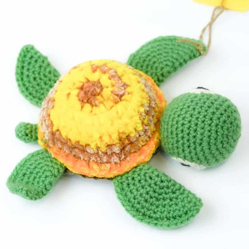 Baby Turtle by Gala Crochet (1) Hand‑crocheted turtle toy made from soft yarn, featuring a round shell in shades of yellow, orange, and light brown, with green crocheted head, flippers, and tail. The turtle rests on a white background, highlighting its bright colours and textured stitching.