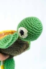 Baby Turtle by Gala Crochet (3) Close‑up of a hand‑crocheted turtle toy made from yarn, showing its green head with a large white and black crocheted eye, and part of its brown shell. The detailed crochet stitches and textures are visible, highlighting the craftsmanship. The background is plain and softly lit to emphasize the turtle’s colour