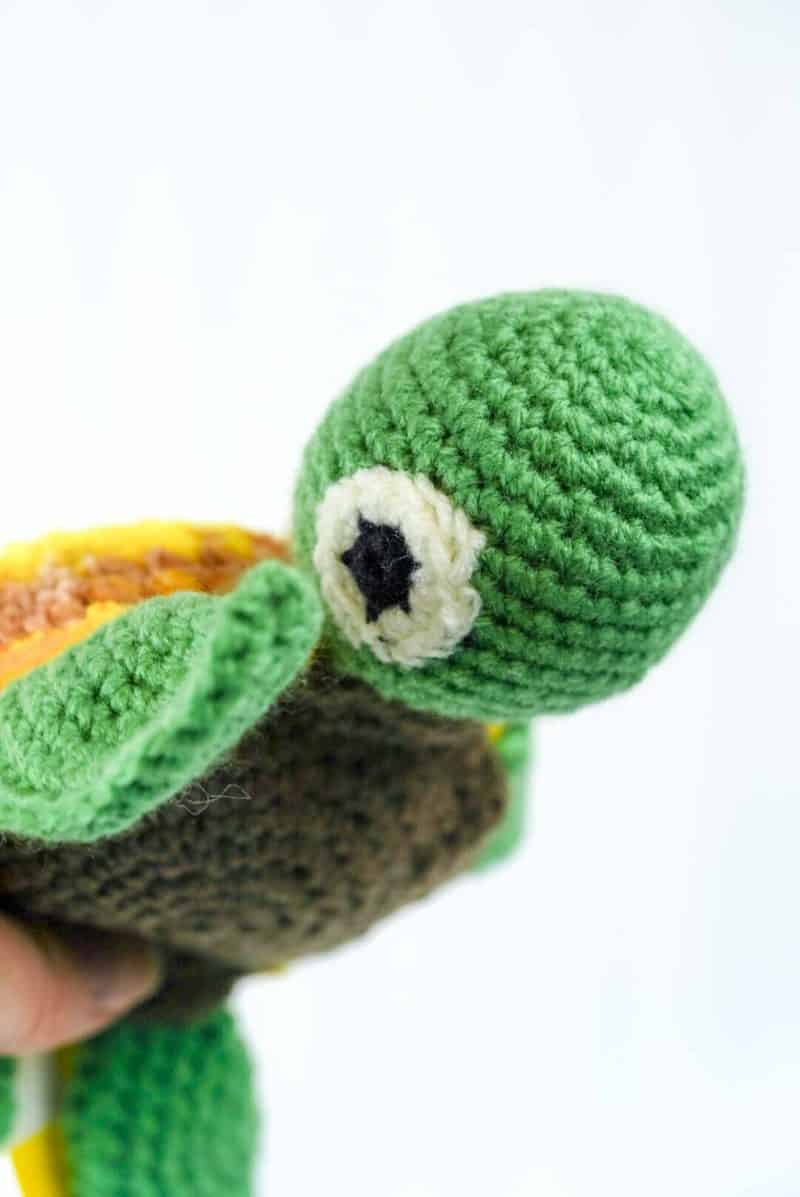 Close‑up of a hand‑crocheted turtle toy made from yarn, showing its green head with a large white and black crocheted eye, and part of its brown shell. The detailed crochet stitches and textures are visible, highlighting the craftsmanship. The background is plain and softly lit to emphasize the turtle’s colour