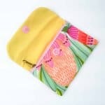 A handmade fabric coin purse with its flap open, revealing a bright yellow interior and a zippered compartment. The outer fabric features a colorful botanical print with stylized flowers and leaves in shades of pink, coral, green, and orange. The purse has a pink snap button on the flap and a gold zipper pull, combining cheerful colors and neat stitching for a vibrant, handcrafted look.