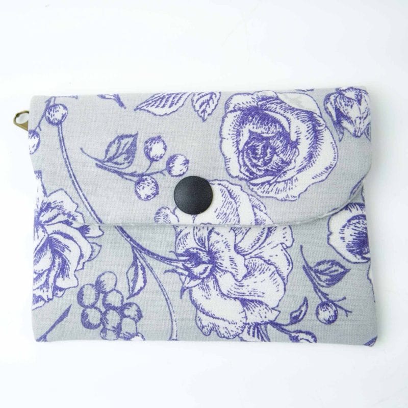 A handmade fabric coin purse with a flap closure secured by a black snap button. The purse features a light grey background with a detailed purple floral print, including roses, leaves, and small berries in a fine line-art style. The fabric has a smooth texture, and a small metal loop is visible on one side, possibly for attaching a strap or keychain. The purse is photographed flat against a plain white surface, showcasing its elegant and vintage-inspired design.