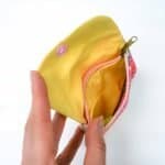 A small handmade fabric coin purse being held open by a hand, showing its bright yellow interior lining. Inside, there’s a zippered compartment with a pink zipper and a gold pull tab. The outer edge of the purse features a colorful floral pattern in shades of pink, coral, and green. The design highlights the purse’s neat stitching, cheerful colors, and practical layout for storing coins or small items.