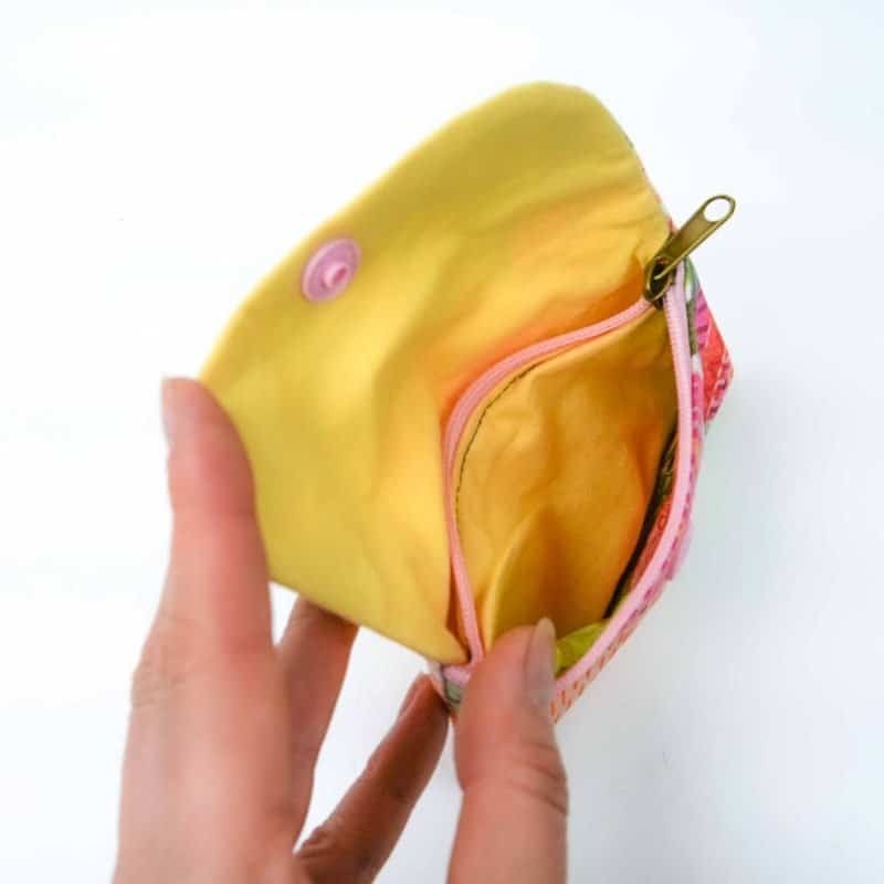 A small handmade fabric coin purse being held open by a hand, showing its bright yellow interior lining. Inside, there’s a zippered compartment with a pink zipper and a gold pull tab. The outer edge of the purse features a colorful floral pattern in shades of pink, coral, and green. The design highlights the purse’s neat stitching, cheerful colors, and practical layout for storing coins or small items.