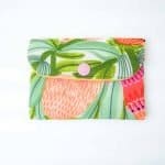 A small handmade fabric coin purse with a flap closure secured by a pink snap button. The purse features a vibrant botanical print with large stylized leaves and flowers in shades of green, coral, pink, and orange. The fabric has a textured weave, and the background is white, highlighting the colorful design. The purse is photographed flat against a plain white surface, showcasing its cheerful, tropical-inspired pattern and neat craftsmanship