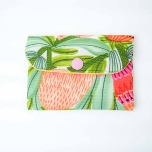 A small handmade fabric coin purse with a flap closure secured by a pink snap button. The purse features a vibrant botanical print with large stylized leaves and flowers in shades of green, coral, pink, and orange. The fabric has a textured weave, and the background is white, highlighting the colorful design. The purse is photographed flat against a plain white surface, showcasing its cheerful, tropical-inspired pattern and neat craftsmanship