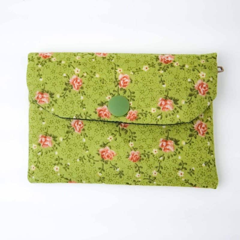 A handmade fabric coin purse with a flap closure secured by a green snap button. The purse features a soft green background patterned with small pink rosebuds and delicate vines, creating a vintage floral look. The fabric has a slightly textured appearance, and a small metal loop is visible on one side, possibly for attaching a strap or keychain. The purse is photographed flat against a plain white surface, highlighting its neat stitching and charming, cottage-style design.