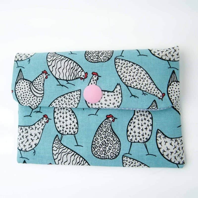 A handmade fabric coin purse with a flap closure secured by a pink snap button. The purse features a light blue background decorated with playful black-and-white chicken illustrations, each with unique geometric patterns such as stripes, dots, and crosses. The chickens have small red combs and thin black legs, creating a whimsical and fun design. The purse is photographed flat against a plain white surface, highlighting its cheerful and quirky pattern.