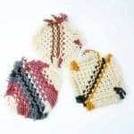 Three handcrafted macramé drink coasters made from thick cotton cord, displayed on a clean white background. Each coaster features a unique woven pattern with fringed edges — one in cream with terracotta and brown stripes, one in cream with mustard and black diagonal stripes, and one in cream with rose, charcoal, and ivory stripes.