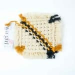 A square macramé drink coaster woven from cream cotton cord with mustard and black diagonal stripes and fringed edges. A WA Girl tag is visible beside the coaster, set against a bright white background.