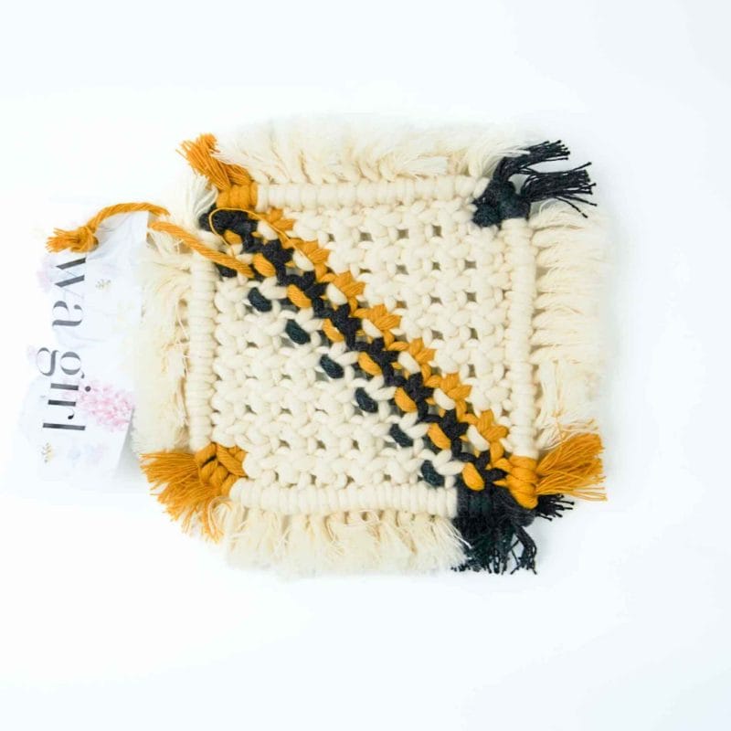 A square macramé drink coaster woven from cream cotton cord with mustard and black diagonal stripes and fringed edges. A WA Girl tag is visible beside the coaster, set against a bright white background.