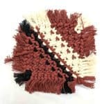 A handcrafted macramé drink coaster woven from cream cotton cord with terracotta, black, and natural accents arranged in a geometric pattern. The coaster has soft fringed edges and is displayed on a clean white background with a WA Girl tag partially visible beside it
