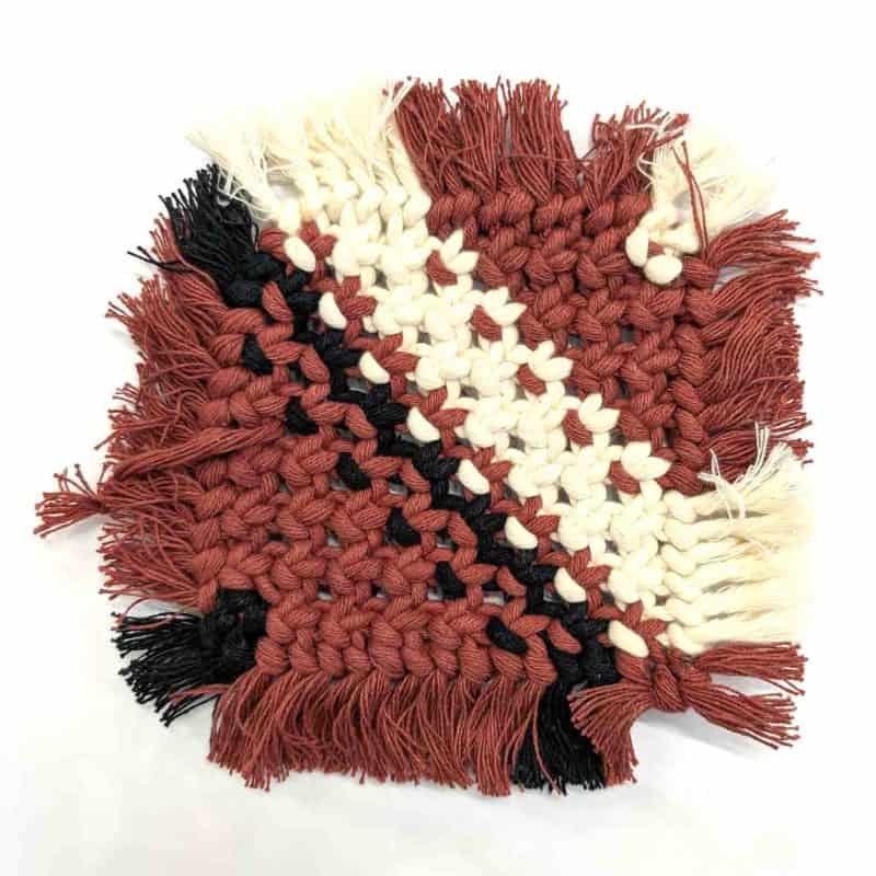 A handcrafted macramé drink coaster woven from cream cotton cord with terracotta, black, and natural accents arranged in a geometric pattern. The coaster has soft fringed edges and is displayed on a clean white background with a WA Girl tag partially visible beside it
