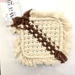 A handcrafted square macramé drink coaster made from thick cream cotton cord with a diagonal stripe of brown woven thread. The coaster features soft fringed edges and a WA Girl tag partially visible beside it, displayed on a clean white background.