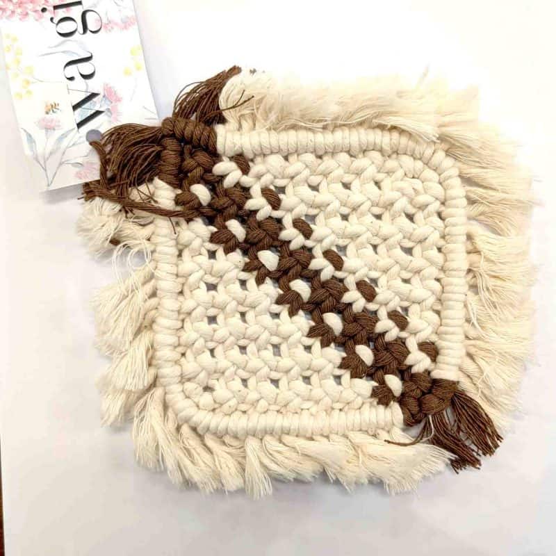 A handcrafted square macramé drink coaster made from thick cream cotton cord with a diagonal stripe of brown woven thread. The coaster features soft fringed edges and a WA Girl tag partially visible beside it, displayed on a clean white background.