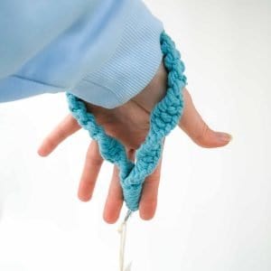 A close‑up of a hand wearing a light blue sleeve, holding a turquoise macramé wristlet keychain made from thick braided cotton cord. The wristlet loops comfortably around the wrist, with a small metal ring and tag hanging below, set against a clean white background.