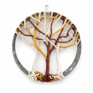 A handcrafted macramé wall hanging depicting a stylized tree of life within a circular metal hoop. The tree is made from cotton cords in cream, golden yellow, and brown tones, with twisted roots at the base and branching limbs that spread across the hoop. The metal ring retains its natural patina, adding rustic character, and the piece is displayed against a clean white background.