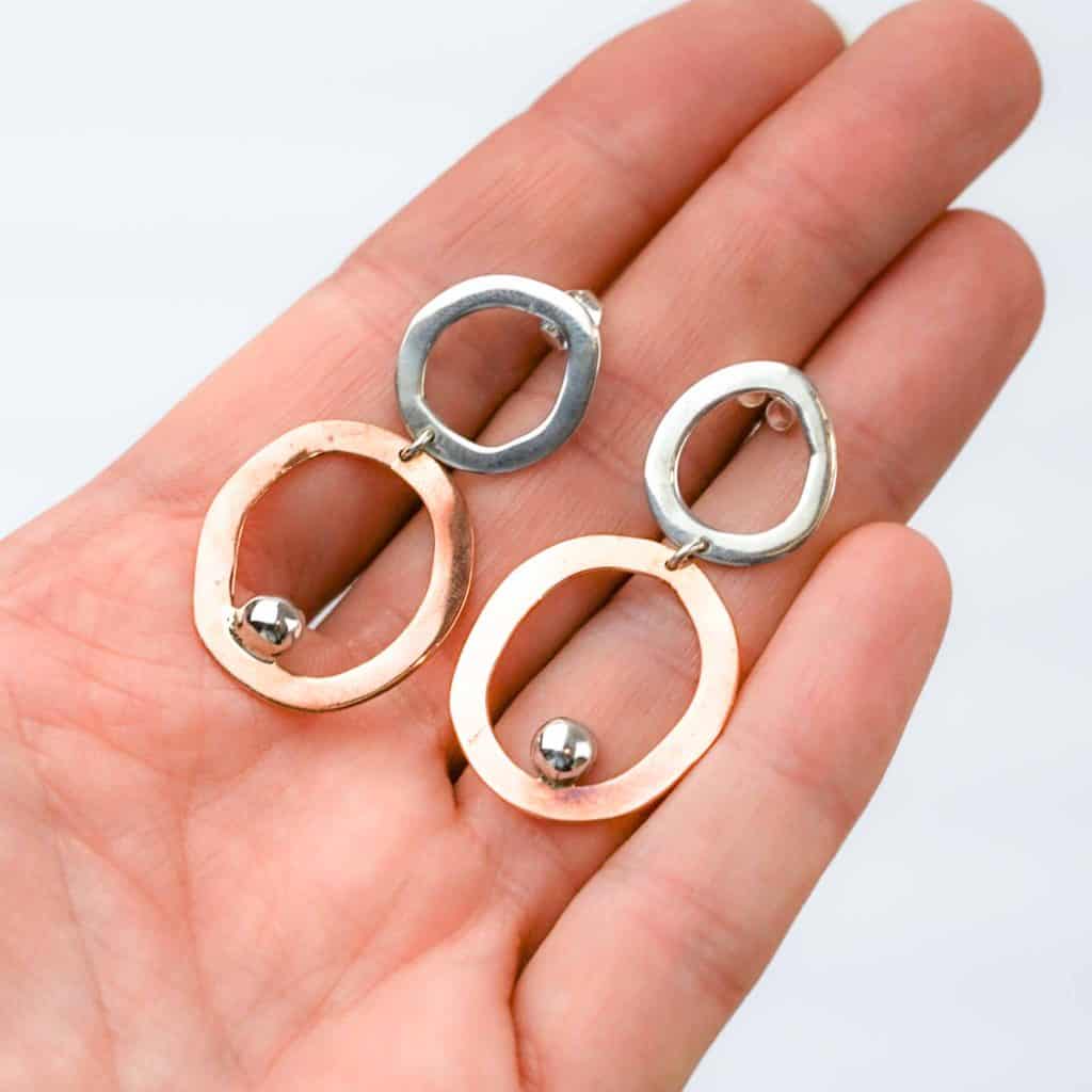 A close-up of a hand holding a pair of handcrafted earrings featuring an abstract, modern design. Each earring has two irregular open hoops — the top hoop in polished sterling silver and the bottom hoop in warm copper — linked together with a small connector. A shiny silver ball embellishment sits near the lower edge of the copper hoop, adding contrast and movement. The earrings have a sculptural, organic shape and are displayed against a soft white background, highlighting their mixed-metal craftsmanship and artistic form.