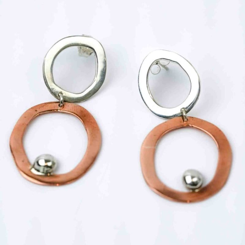 A pair of handcrafted earrings displayed against a plain white background. Each earring features two irregular open hoops — the upper hoop in polished sterling silver and the lower hoop in warm copper — connected by a small link. A shiny silver ball embellishment sits near the bottom edge of the copper hoop, adding contrast and movement. The earrings have a sculptural, organic design that highlights their mixed-metal craftsmanship and modern artistic style.