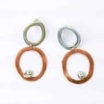 Sterling silver copper abstract duo hoop ER with SS ball embellishment (4) A pair of handcrafted earrings featuring an abstract, modern design. Each earring consists of two irregular open hoops β the upper hoop in polished sterling silver and the lower hoop in warm copper β linked together with a small connector. A shiny silver ball embellishment rests near the bottom edge of the copper hoop, adding contrast and movement. The earrings have a sculptural, organic shape and are displayed against a plain white background, highlighting their mixed-metal craftsmanship and artistic form.