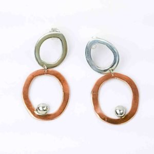 A pair of handcrafted earrings featuring an abstract, modern design. Each earring consists of two irregular open hoops — the upper hoop in polished sterling silver and the lower hoop in warm copper — linked together with a small connector. A shiny silver ball embellishment rests near the bottom edge of the copper hoop, adding contrast and movement. The earrings have a sculptural, organic shape and are displayed against a plain white background, highlighting their mixed-metal craftsmanship and artistic form.