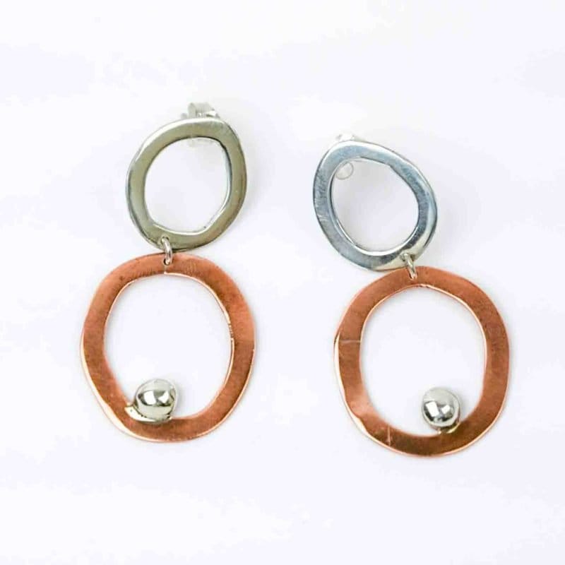 Sterling silver copper abstract duo hoop ER with SS ball embellishment (4) A pair of handcrafted earrings featuring an abstract, modern design. Each earring consists of two irregular open hoops β the upper hoop in polished sterling silver and the lower hoop in warm copper β linked together with a small connector. A shiny silver ball embellishment rests near the bottom edge of the copper hoop, adding contrast and movement. The earrings have a sculptural, organic shape and are displayed against a plain white background, highlighting their mixed-metal craftsmanship and artistic form.