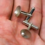 A close-up image of a hand holding a pair of sterling silver cufflinks. Each cufflink features a concave circular face with a smooth satin finish and a single white freshwater pearl set near the center. The silver has a polished sheen, and the cufflinks’ cylindrical bars and hinges are visible, showcasing their fine craftsmanship. The warm skin tone of the hand provides a soft background that highlights the elegant contrast between the lustrous pearl and the metallic silver.