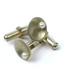 A close-up image of a pair of handcrafted sterling silver cufflinks displayed on a white background. Each cufflink features a concave circular face with a smooth satin finish and a single white freshwater pearl set near the center. The silver has a soft matte texture, and the cufflinks are arranged to show both the decorative face and the polished swivel bar mechanism. The composition highlights the elegant contrast between the lustrous pearl and the brushed silver surface.