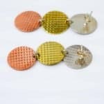 Trio textured cppper s silver brass disc earrings (1) A pair of handcrafted earrings featuring three linked circular discs in metallic tones of copper, brass, and silver. Each disc has a subtle grid-textured surface that reflects light, creating a shimmering effect. The discs are connected in a vertical arrangement, forming a modern, geometric design. The earrings have stud-style posts with butterfly backs, and the mixed-metal palette gives them a warm, contemporary look against a plain white background.
