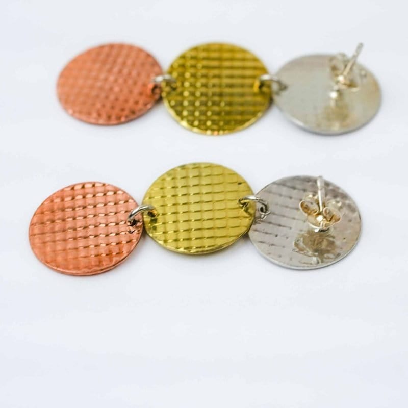 A pair of handcrafted earrings featuring three linked circular discs in metallic tones of copper, brass, and silver. Each disc has a subtle grid-textured surface that reflects light, creating a shimmering effect. The discs are connected in a vertical arrangement, forming a modern, geometric design. The earrings have stud-style posts with butterfly backs, and the mixed-metal palette gives them a warm, contemporary look against a plain white background.