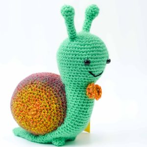 A handmade crochet snail toy crafted from yarn, featuring a cheerful design. The snail has a bright green body with two antennae, black bead eyes, and a stitched smile. Its shell is round and multicoloured, blending shades of red, orange, and yellow in a gradient pattern. A small orange crochet bow sits at the front of its neck. The background is plain white, highlighting the vibrant colours and detailed stitching of the snail.