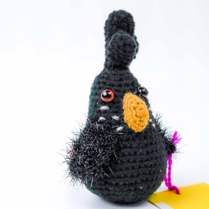 This image shows a hand‑crocheted black cockatoo toy made from yarn. The bird has a rounded body with a tall crest on its head, a bright yellow crocheted beak, and shiny red plastic eyes. Its wings are made from fuzzy black yarn with metallic threads that give a textured, feathery look. A short piece of purple yarn extends from one side, and a yellow tag lies beside it on a white background.