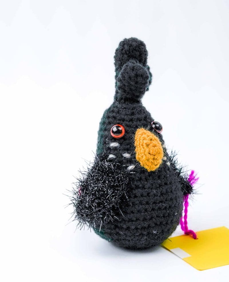 hand‑crocheted black cockatoo toy by Gala Crochet (3) This image shows a hand‑crocheted black cockatoo toy made from yarn. The bird has a rounded body with a tall crest on its head, a bright yellow crocheted beak, and shiny red plastic eyes. Its wings are made from fuzzy black yarn with metallic threads that give a textured, feathery look. A short piece of purple yarn extends from one side, and a yellow tag lies beside it on a white background.