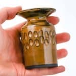 A small handcrafted ceramic vase made from black clay, shown being held in a person’s hand for scale. The vase has a glossy mustard glaze with darker accents and a flared rim. Around the middle, a decorative band of carved oval patterns adds texture and visual interest. The glaze reflects light softly, highlighting the artisanal craftsmanship and warm earthy tones against a plain white background.