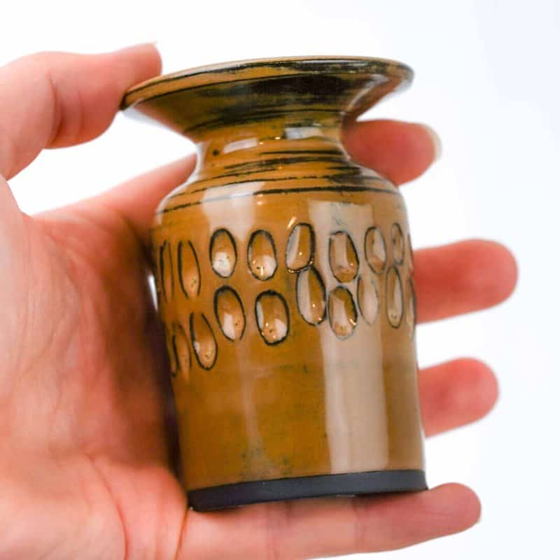 A small handcrafted ceramic vase made from black clay, shown being held in a person’s hand for scale. The vase has a glossy mustard glaze with darker accents and a flared rim. Around the middle, a decorative band of carved oval patterns adds texture and visual interest. The glaze reflects light softly, highlighting the artisanal craftsmanship and warm earthy tones against a plain white background.
