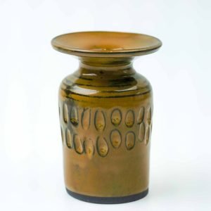 A handcrafted ceramic vase made from black clay with a glossy mustard glaze. The vase has a cylindrical body that narrows at the neck and flares into a wide, flat rim. Around the middle section, a series of carved oval patterns create a textured decorative band. The glaze shows subtle tonal variations, giving the piece a warm, artisanal finish against a plain white background.