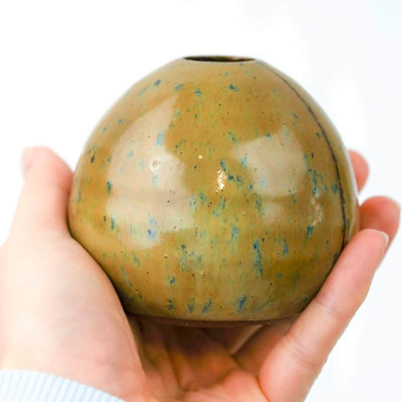A small, handcrafted round vase made from Klamath clay, featuring a smooth glossy surface with an ochre-brown base and subtle blue-green speckles. The vase has a narrow opening at the top and a gentle stripe detail, finished with an opal lustre glaze held in the palm of a hand.