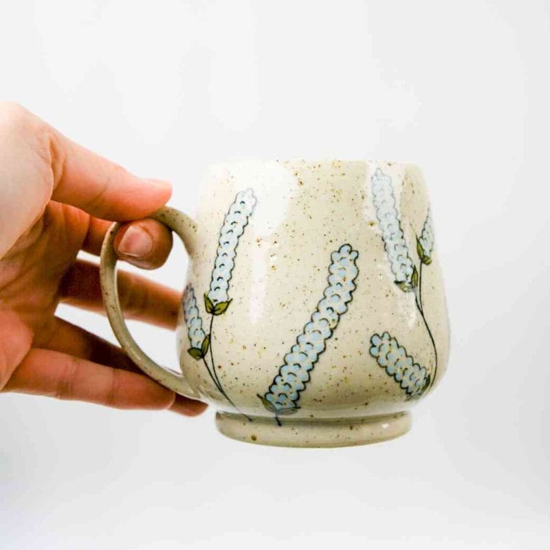 A hand holding a handcrafted ceramic mug against a plain white background. The mug has a rounded shape with a speckled beige glaze and a sturdy handle. Its surface is decorated with hand-painted baby blue lupin flowers featuring green stems and leaves, arranged in a delicate repeating pattern. The design highlights the mug’s artisanal craftsmanship and botanical elegance.
