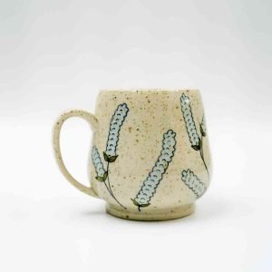A handcrafted ceramic mug with a rounded shape and a single handle, displayed against a plain white background. The mug has a speckled beige glaze and is decorated with hand-painted baby blue lupin flowers featuring green stems and leaves. The floral design wraps around the mug, giving it a delicate, botanical look. The piece has a smooth finish and a slightly raised base, highlighting its artisan craftsmanship.