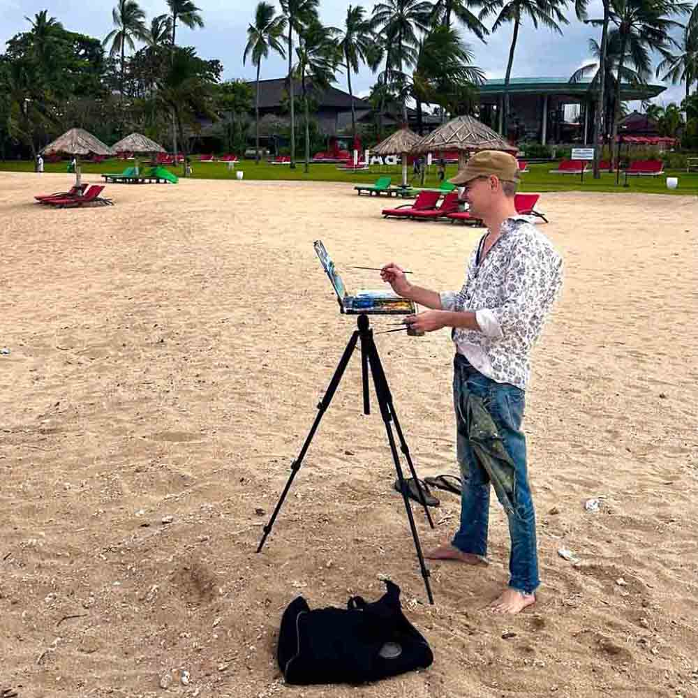 Artist Ben Sherar stands barefoot on a sandy beach in Nusa Dua, wearing a cap and patterned shirt, while painting on a canvas attached to a portable tripod easel with palm trees and beach loungers in the background.