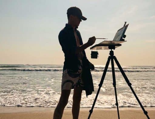 Silhouette of a man wearing a baseball cap and shorts, painting on an easel at Kuta Beach, Bali, during a bright, sunny day with ocean waves crashing in the background.