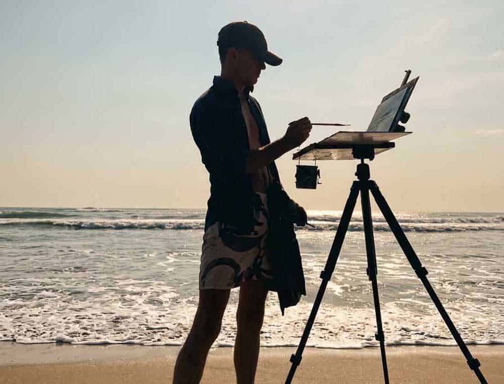 Silhouette of a man wearing a baseball cap and shorts, painting on an easel at Kuta Beach, Bali, during a bright, sunny day with ocean waves crashing in the background.