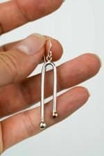 A close-up of a hand holding a single earring made from polished sterling silver. The earring has a sleek, elongated U-shape design with two rounded ends — one finished in silver and the other in copper — creating a subtle two-tone effect. The piece hangs from a simple silver hook, and the background is plain and light, highlighting the reflective shine of the metal.