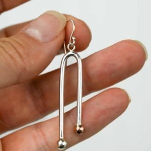 A close-up of a hand holding a single earring made from polished sterling silver. The earring has a sleek, elongated U-shape design with two rounded ends — one finished in silver and the other in copper — creating a subtle two-tone effect. The piece hangs from a simple silver hook, and the background is plain and light, highlighting the reflective shine of the metal.