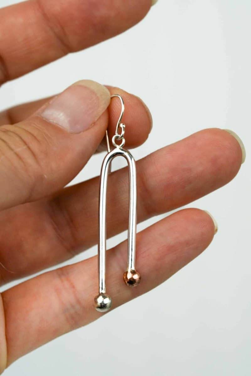 A close-up of a hand holding a single earring made from polished sterling silver. The earring has a sleek, elongated U-shape design with two rounded ends — one finished in silver and the other in copper — creating a subtle two-tone effect. The piece hangs from a simple silver hook, and the background is plain and light, highlighting the reflective shine of the metal.