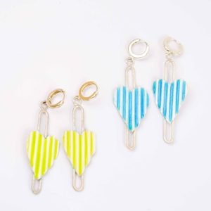 Two pairs of corrugated heart earrings are displayed on a white background. Each earring features a vertically striped enamel heart suspended within a slender sterling silver frame. One pair has bright lime green and white stripes with gold hoops, while the other pair has blue and white stripes with silver hoops. The design is modern and playful, showcasing handcrafted detail and vibrant colour contrast.
